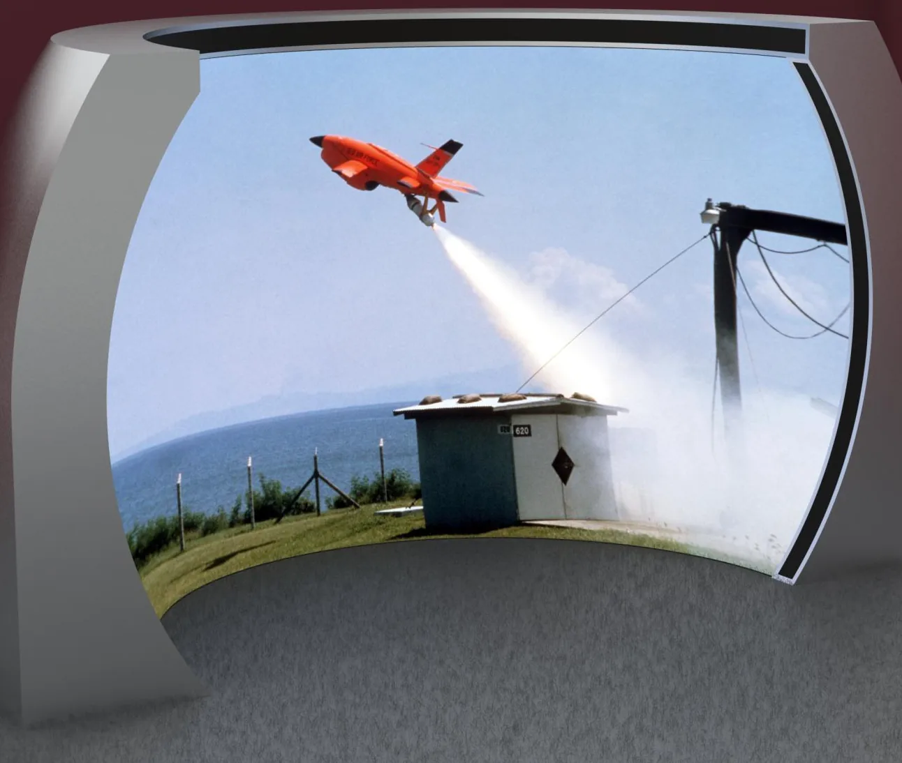 A forward-view immersive display standing on its own: one continuous curved screen wrapped through 180 degrees, carried on a shell that closes behind it, showing a coastal test range with an orange aircraft in flight.